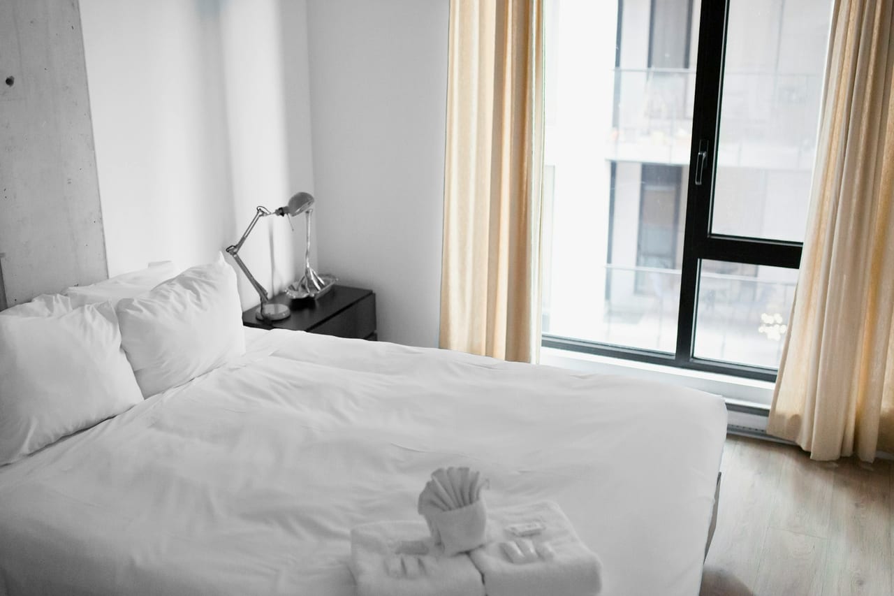 A neatly made hotel bed beside a bright window