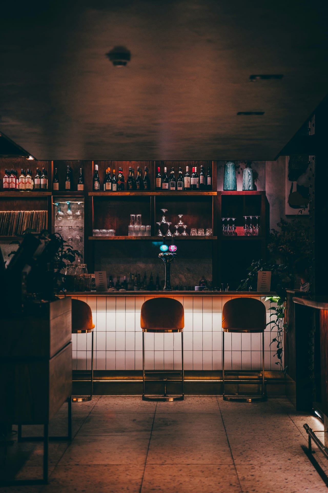 A dimly lit bar with stools and back-bar shelves